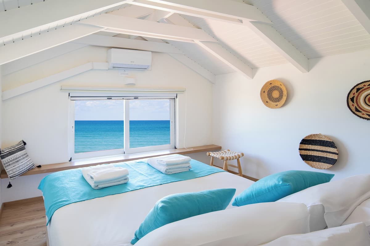 A bedroom with a bed, a window with a view of the sea, a ceiling fan, and a small stool.