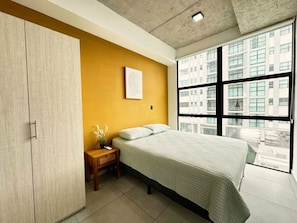 A bedroom with a bed, a nightstand, a wall-mounted picture, and a window with a view of buildings. - Big & Modern Apartment with Great Amenities: Children’s Playroom, Pool & Gym! (Nuevo Cuscatlan)