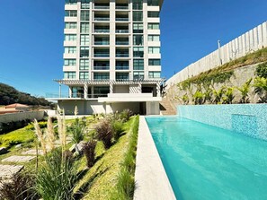 A modern hotel with a swimming pool, a landscaped garden, and a view of the surrounding area. - Big & Modern Apartment with Great Amenities: Children’s Playroom, Pool & Gym! (Nuevo Cuscatlan)