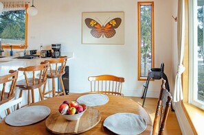 Un coin repas avec une table en bois, des chaises, un saladier rempli de fruits, une machine à café et un tableau représentant un papillon accroché au mur.