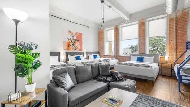 Une chambre d’hôtel moderne avec un canapé gris, un lit avec un oreiller bleu, une grande fenêtre et une plante.