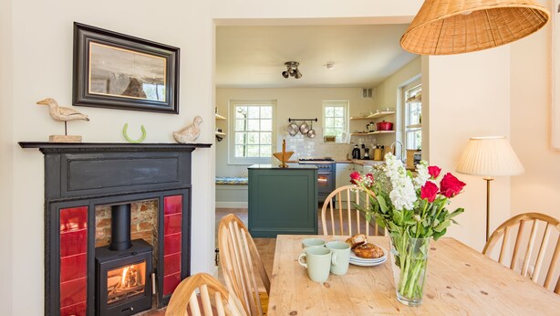 Ein Essbereich mit Kamin, Holzstühlen, einem Tisch mit Blumenvase und einer Küche im Hintergrund.