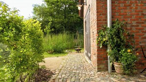 A brick building with a cobblestone pathway leading to a garden area with greenery and a metal fence. - Oasis of peace near the lake (Langen Jarchow)