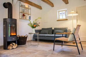 Ein Wohnzimmer mit Kaminofen, einer Couch, einem Korbsessel, einem Tisch mit Blumenvase und einer Lampe.