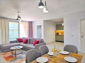 Ein modernes Wohnzimmer mit grauem Sofa, einem Esstisch für zwei Personen und einem durch eine offene Tür einsehbaren Küchenbereich.
