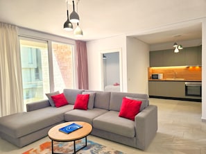 Ein modernes Wohnzimmer mit einem grauen Ecksofa, roten Kissen, einem runden Couchtisch und Blick auf die Stadt durch große Fenster.