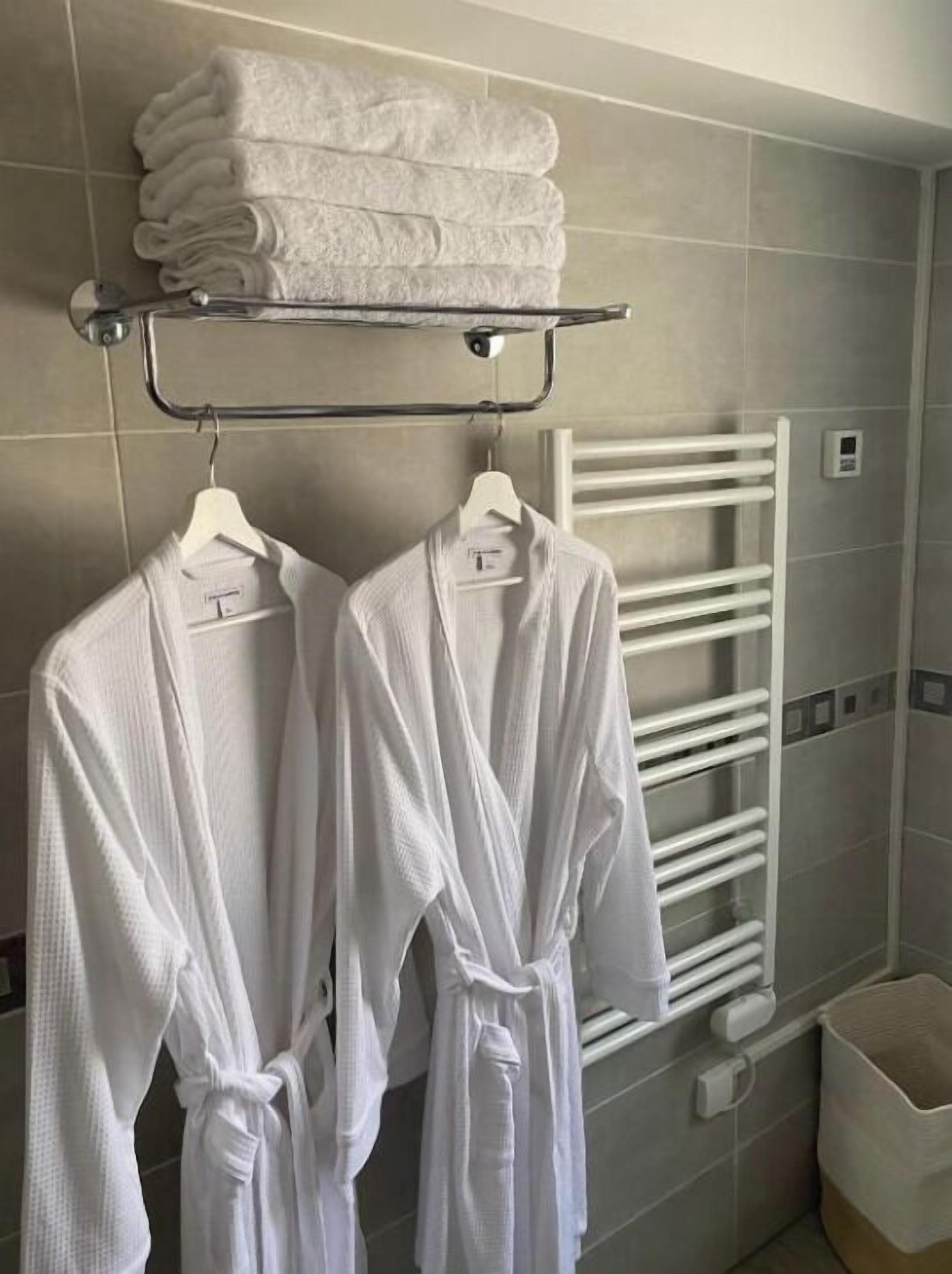 A bathroom with two white robes hanging on a rack, a towel rack with folded towels, a white towel warmer, and a trash bin.