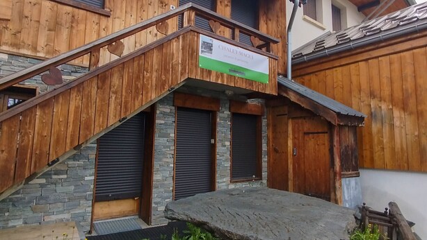 Een houten chalet met een stenen trap, een bordje met de tekst 'Locatie & personen' en een stenen bankje op de voorgrond.