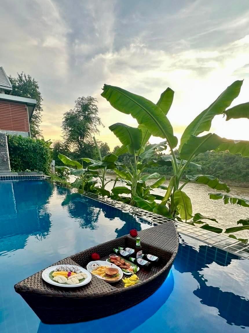 A poolside setting with a dining setup on a boat, surrounded by lush greenery and a view of a river.