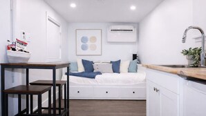 A compact room with a bed, a small table, a sink, and an air conditioning unit.