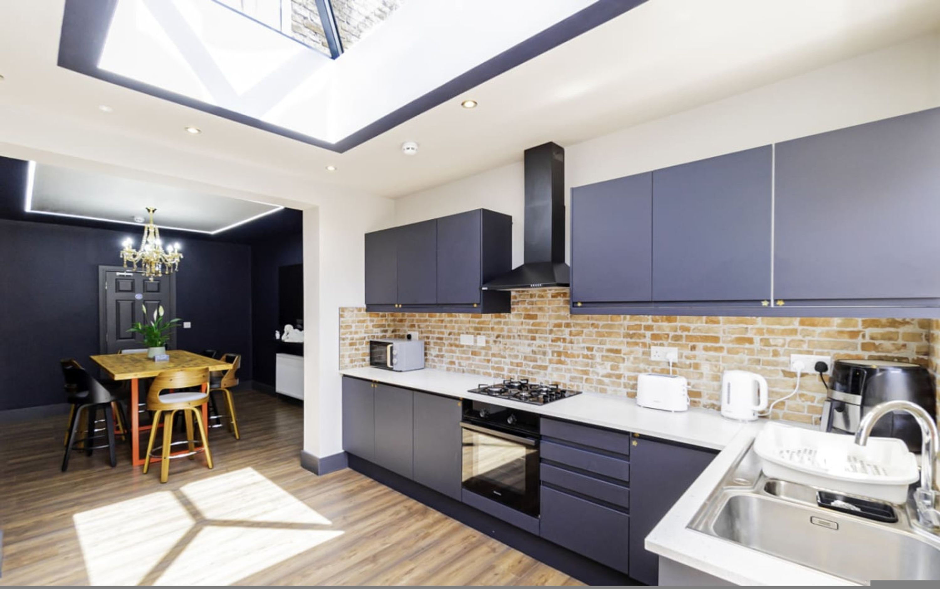 A modern kitchen with dark cabinets, a brick backsplash, a skylight, and a dining area with a wooden table and chairs.