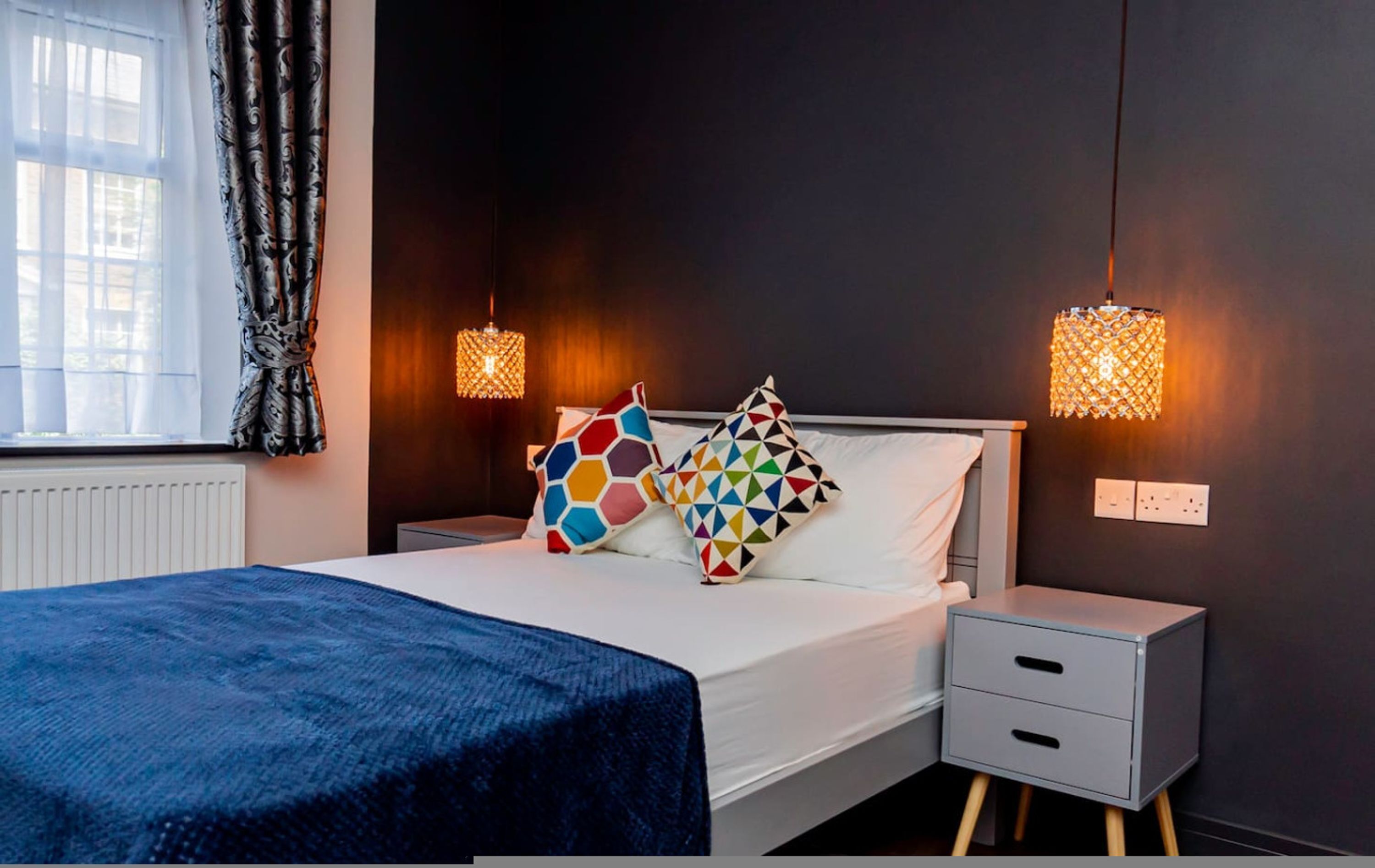A bedroom with a bed, two bedside tables, a window with curtains, and two wall-mounted lamps.
