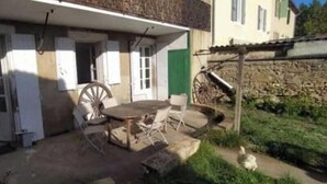 Un edificio rústico con un patio que tiene una mesa, sillas, una rueda de madera y un muro de piedra.