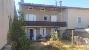 Un edificio de dos pisos con techo plano, una puerta principal y un pequeño jardín con un banco y una fuente de agua.