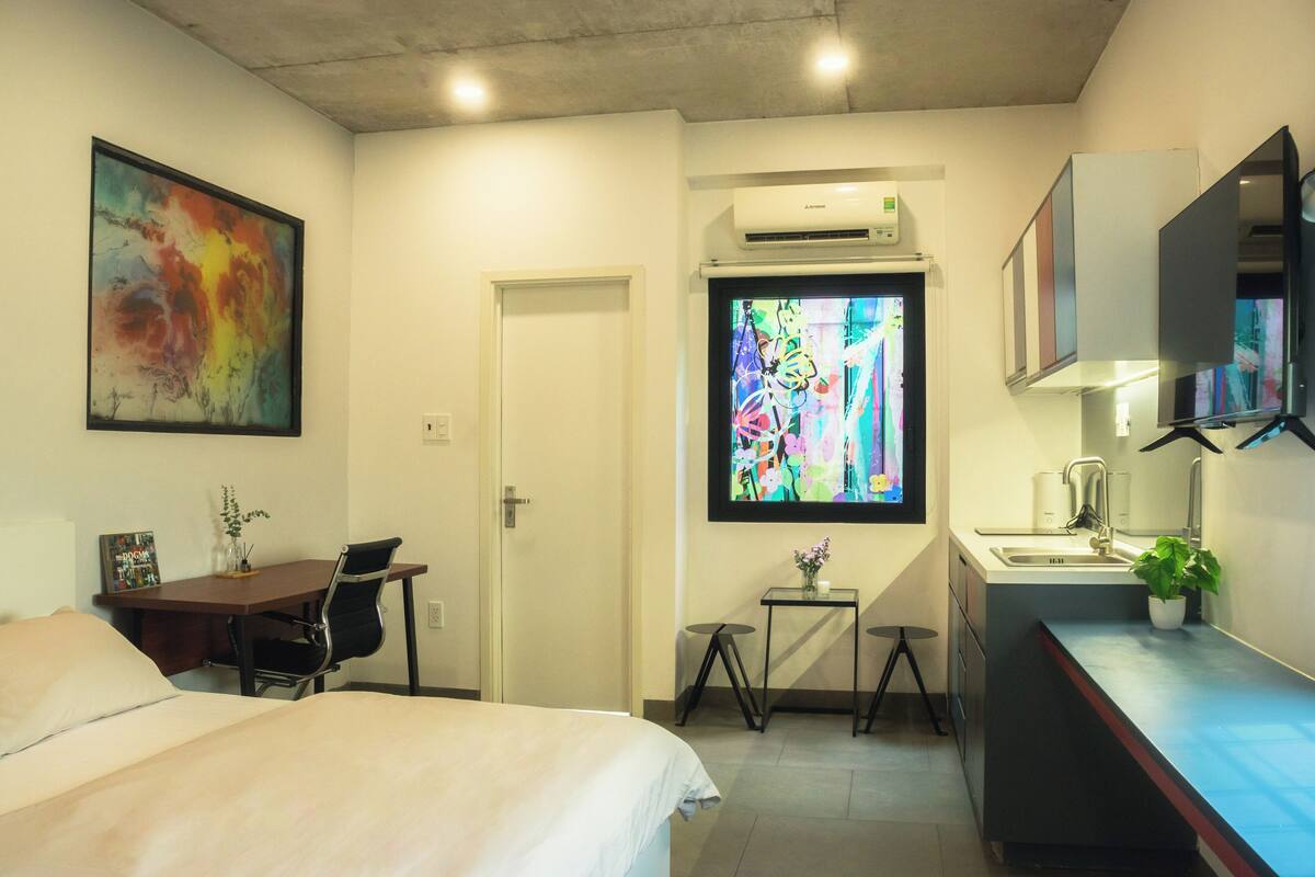 A hotel room with a bed, desk, chair, small table, air conditioning unit, and a large window with a colorful view.