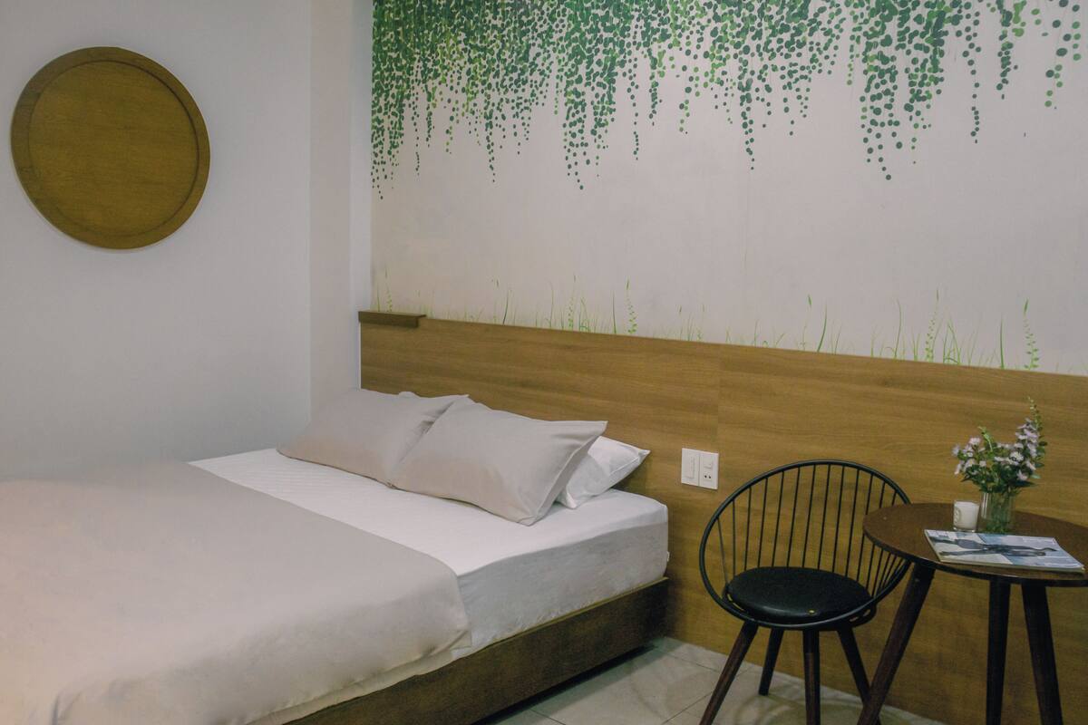 A bed with white linens, a wooden headboard, a wall-mounted round mirror, a black chair, and a small table with a vase of flowers.