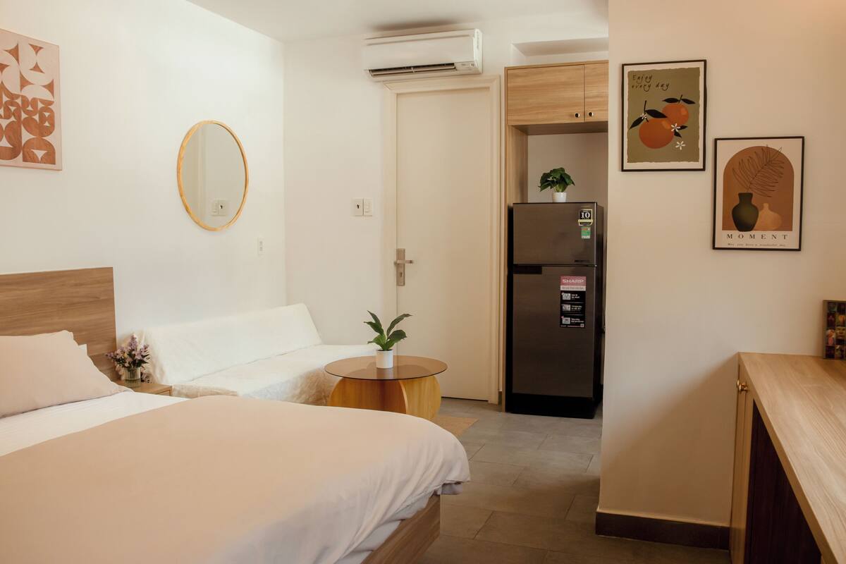 A hotel room with a bed, a round mirror, a small wooden table with a plant, a refrigerator, and a door leading to another room.