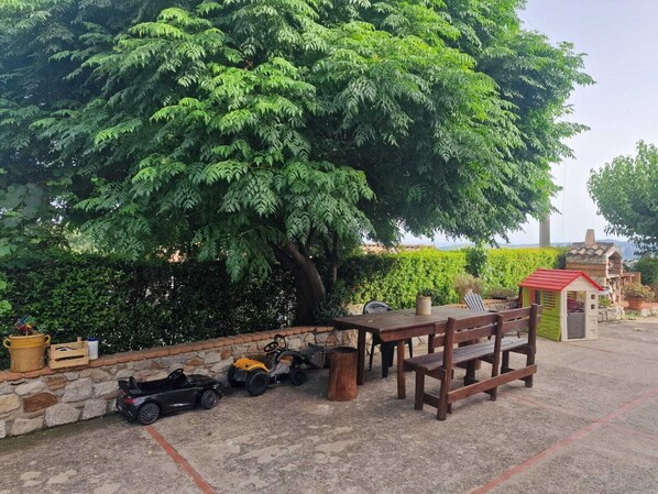 A patio area with a large tree, a wooden bench, a small play structure, and a stone wall.