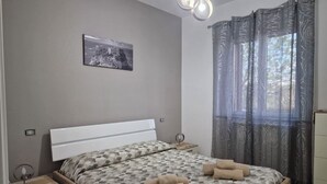 A bedroom with a bed, a nightstand, a wall-mounted light, a window with curtains, and a picture on the wall. - 🦋the village villa 🦋 (Santa Caterina dello Ionio)