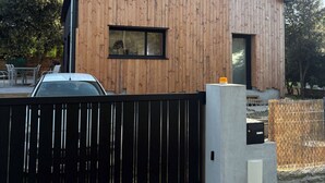 Ein modernes Holzgebäude mit schwarzem Metallzaun, einem kleinen Fenster und einer Betonkonstruktion nahe dem Eingang.