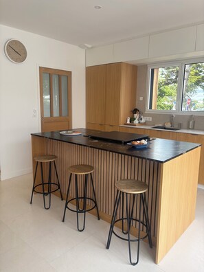 A modern kitchen with a central island, wooden cabinets, and a window with a view. - Maison familiale à l'Ile Tudy - At the end of the street, it's the sea! (Île-Tudy)