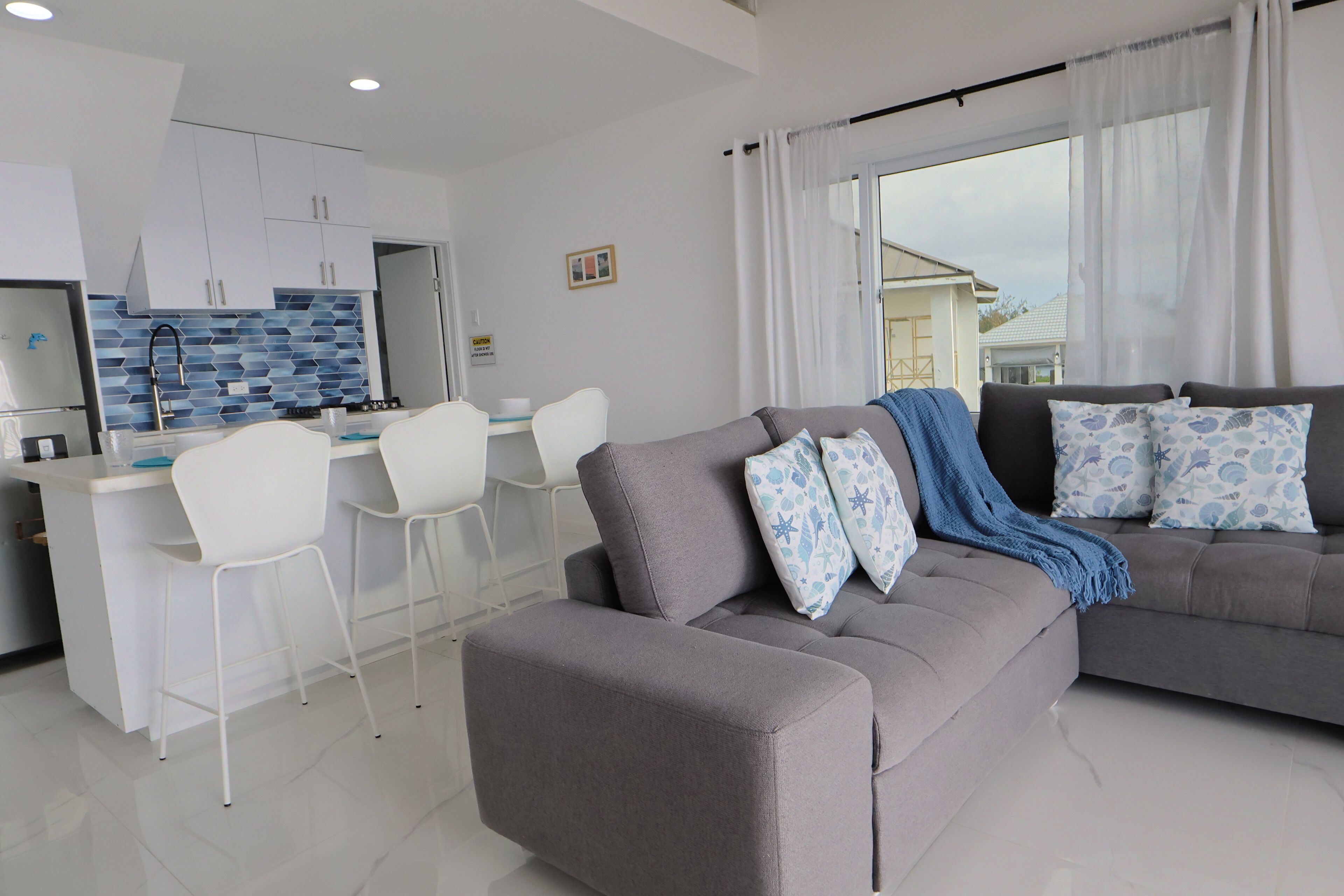 Un soggiorno moderno con un divano grigio, sedie bianche, una zona cucina e vista attraverso un'ampia finestra.