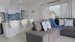 Een moderne woonruimte met een grijze bank, witte stoelen, een keuken en een uitzicht door een groot raam.