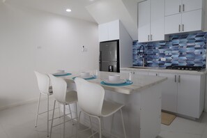A modern kitchen with white cabinetry, a stainless steel refrigerator, a blue-tiled backsplash, and a small island with bar stools.