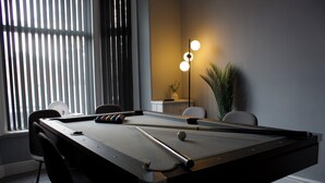 Une table de billard avec des chaises, une lampe et une plante dans une pièce dotée d’une fenêtre et de stores.