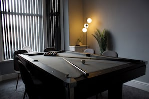Ein Billardtisch mit Stühlen, einer Lampe und einer Pflanze in einem Raum mit Fenster und Jalousien.