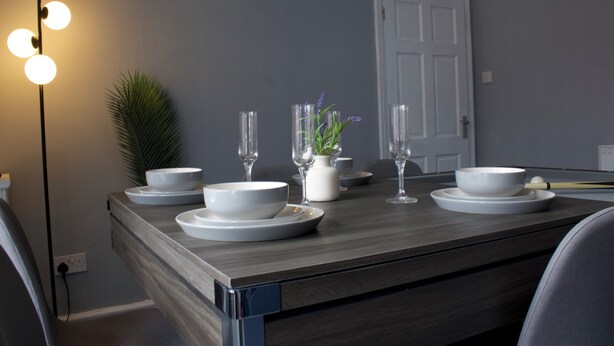Un coin repas avec une table en bois dressée pour quatre personnes, de la verrerie transparente et une plante en pot.