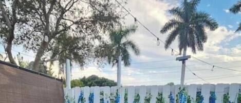 Un jardín trasero con césped artificial, mobiliario de mimbre con cojines azules, una planta en maceta y una cerca blanca con enredaderas azules colgantes.