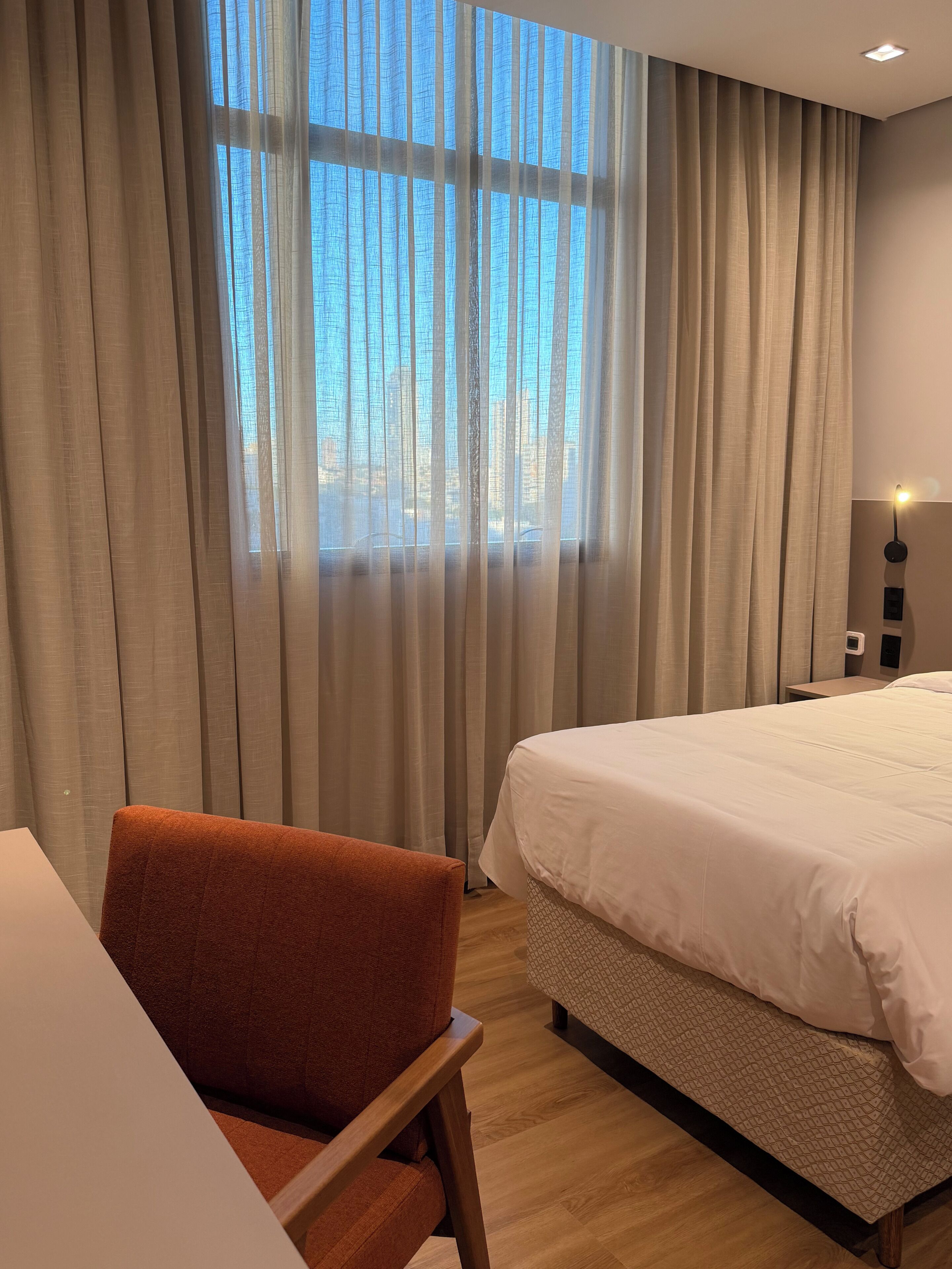 Habitación de hotel con cama, una silla, un escritorio y un amplio ventanal con cortinas.