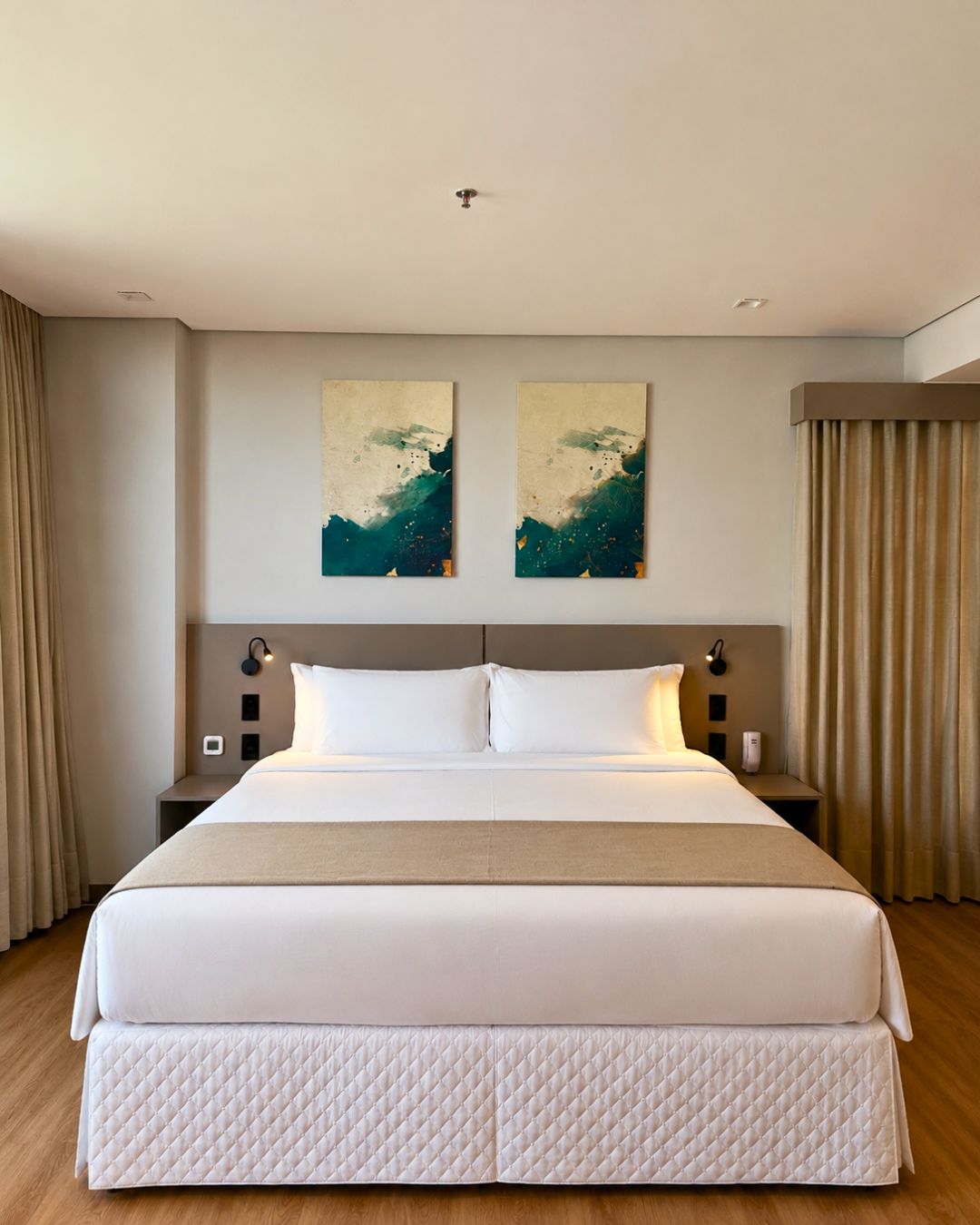 Habitación de hotel con una cama grande, dos obras de arte colgadas en la pared, una mesita de noche con lámpara y una cortina a un lado.