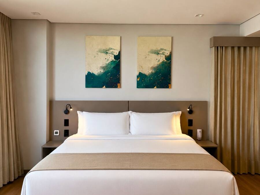 Habitación de hotel con una cama grande, dos obras de arte colgadas en la pared, una mesita de noche con lámpara y una cortina a un lado.