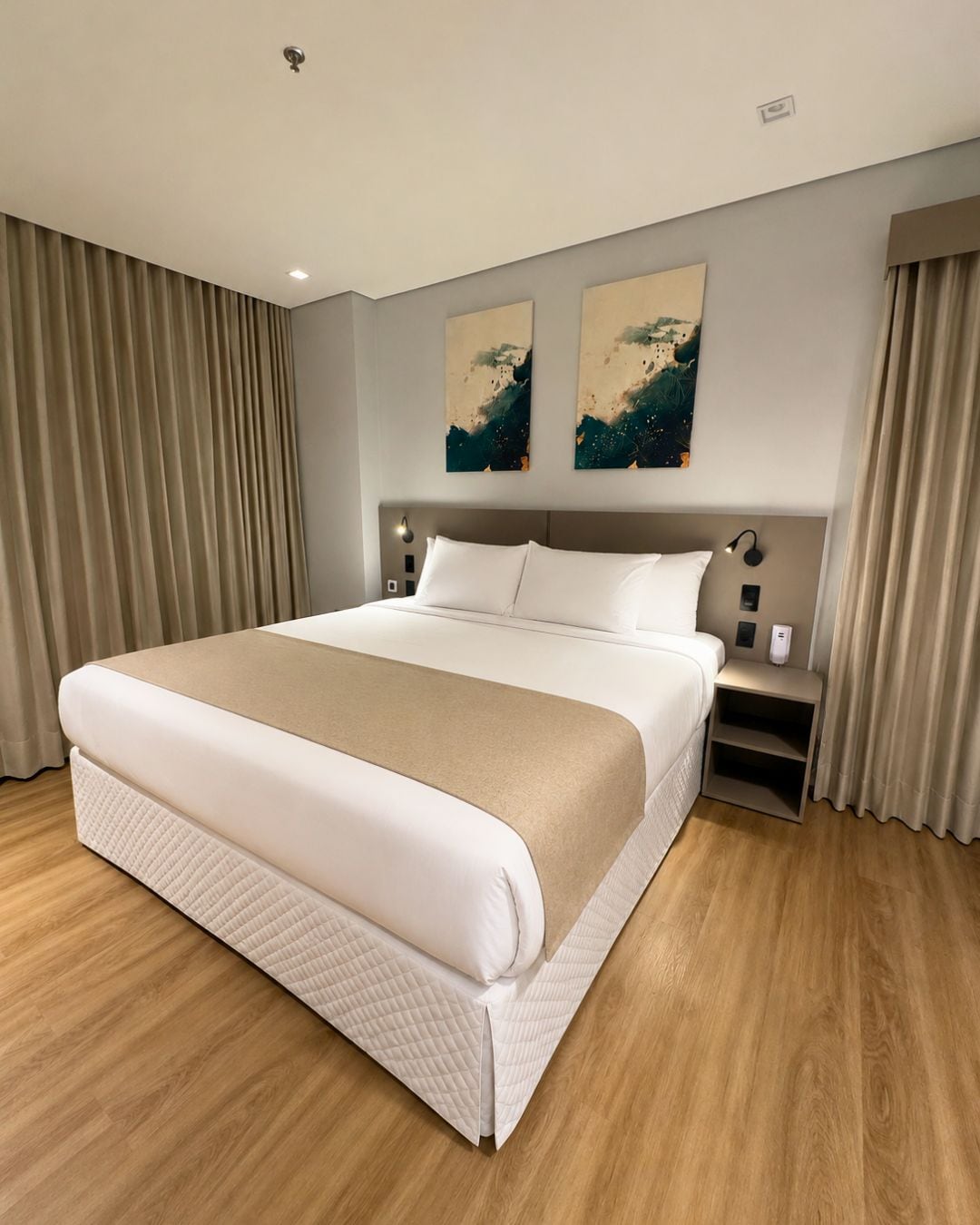 A hotel room with a large bed, a bedside table, a nightstand, and two abstract paintings on the wall.