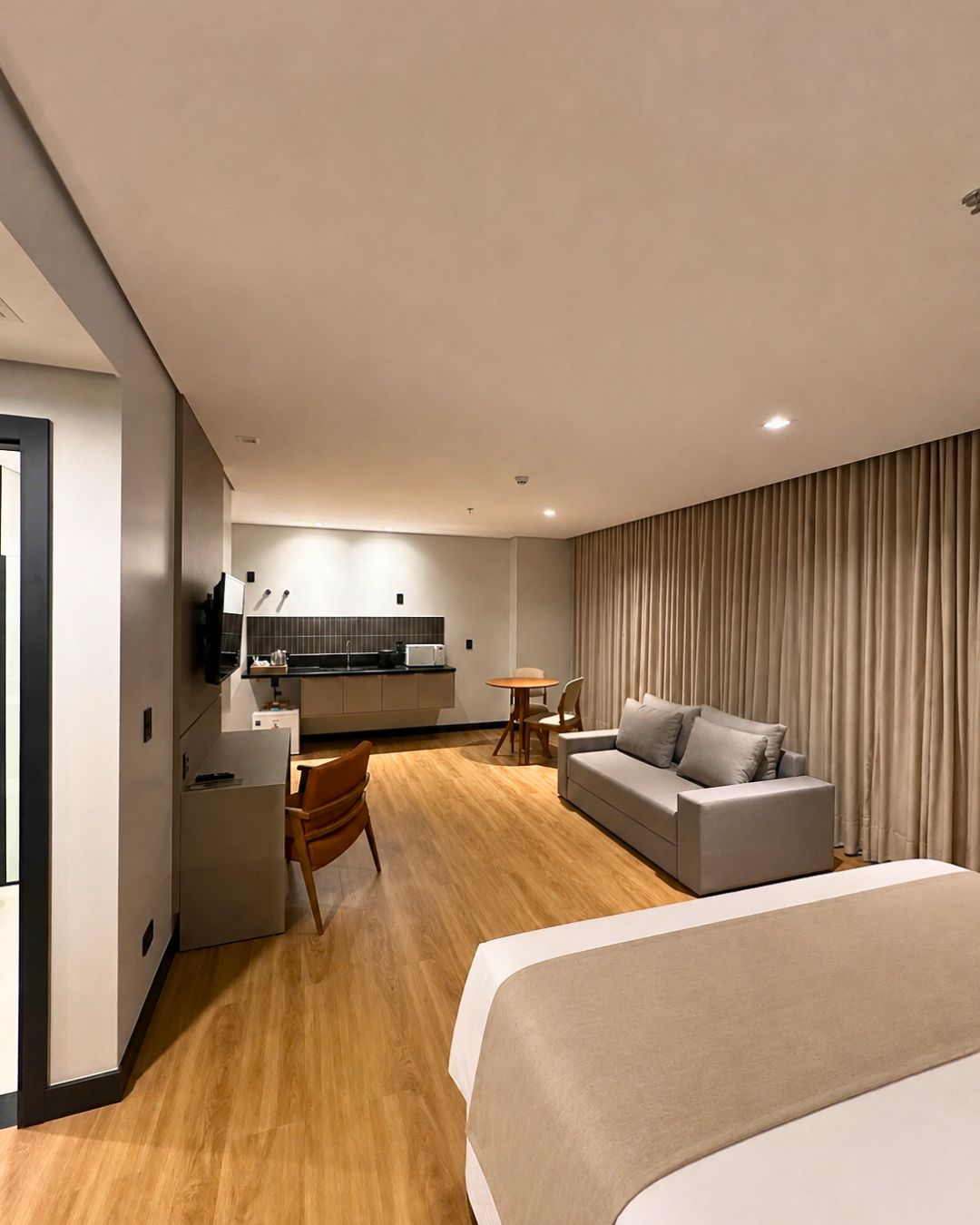 A modern hotel room with a large bed, a sofa, a dining area, and a kitchenette.