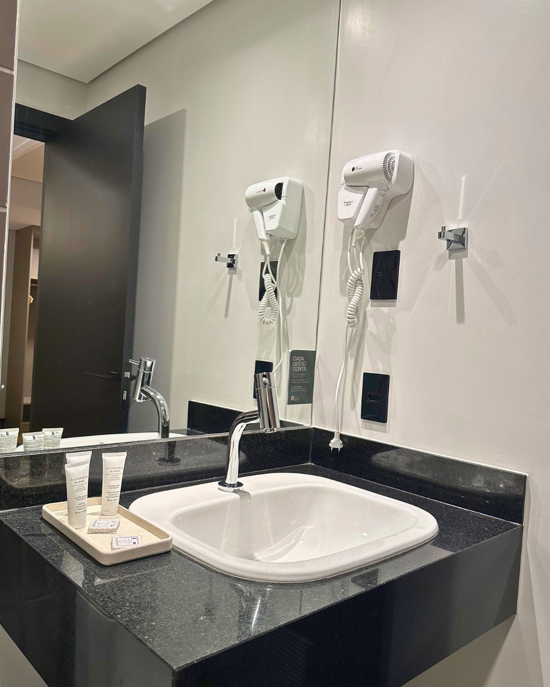 Un baño de hotel con un lavamanos blanco, encimera negra y dos secadores de pelo fijados en la pared.