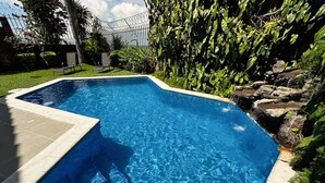 Una piscina rectangular con agua azul cristalina, rodeada de un borde de piedra y abundante vegetación.