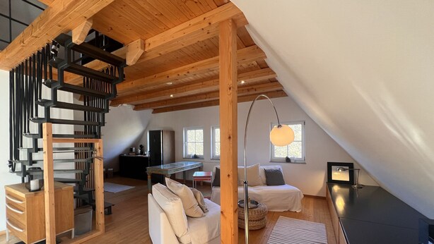 Et loftstudie med en trætrappe, en sofa, et spisebord og en hængende lampe.