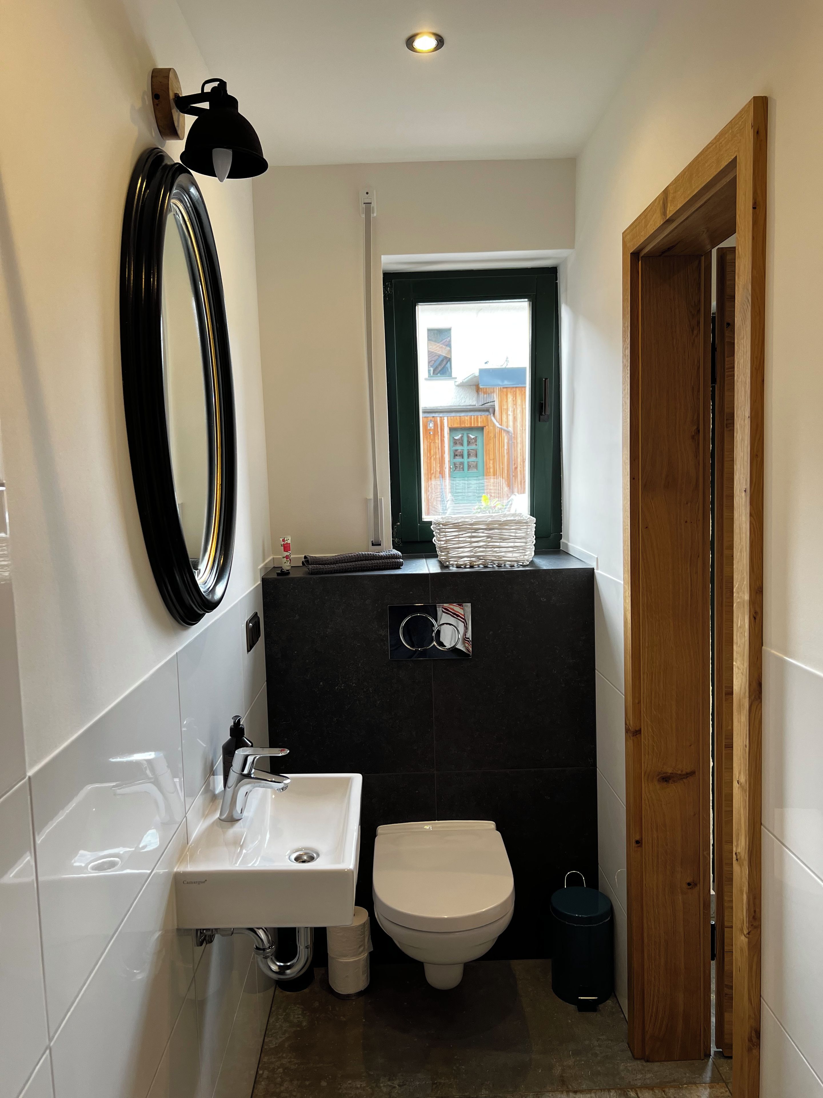 A compact bathroom with a white sink, a toilet, a mirror, and a window with a view of the outside.