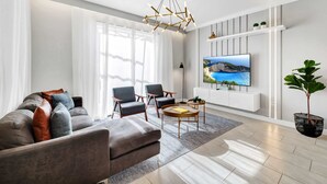 Ein modernes Wohnzimmer mit Sofa, Sesseln, Couchtisch, Fernseher an der Wand und einem großen Fenster.