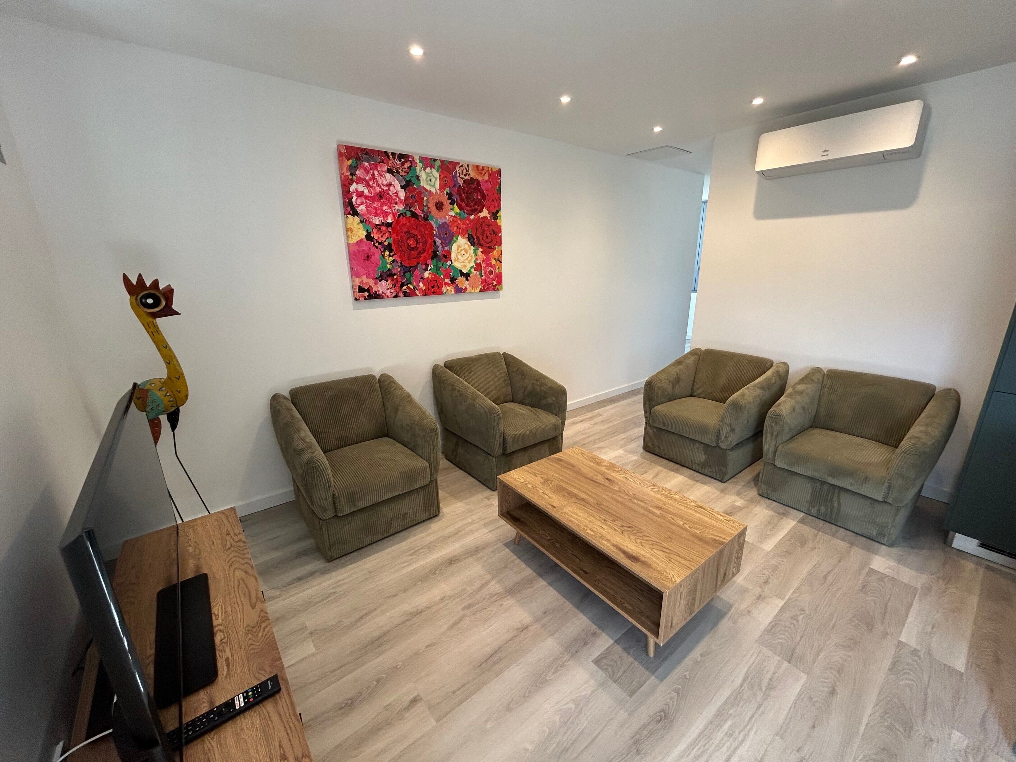 Un salon moderne avec quatre canapés verts, une table basse en bois, un tableau floral aux couleurs vives et une sculpture murale.