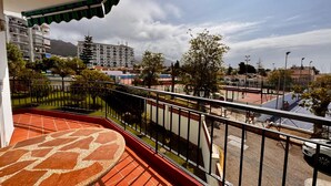 Un balcón con piso de patrón rojo, baranda de metal y vista a una cancha deportiva y edificios al fondo.