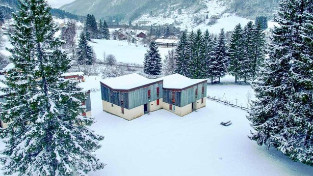 A snow-covered building with a flat roof, surrounded by snow-laden trees and a mountainous backdrop. - 2-bedroom Home in Monts Jura (Lelex)