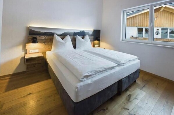 Une chambre à coucher comprenant un lit, des tables de chevet, une tête de lit ornée d’un tableau représentant un paysage, une fenêtre donnant sur un immeuble et un plancher en bois.
