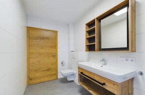 Une salle de bain avec une porte en bois, des carreaux blancs, un lavabo blanc et un miroir avec des étagères.