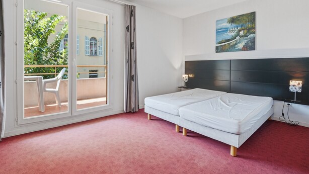 Une chambre d’hôtel avec un lit, un tableau, une table de chevet, une lampe et une vue sur un balcon avec des chaises.