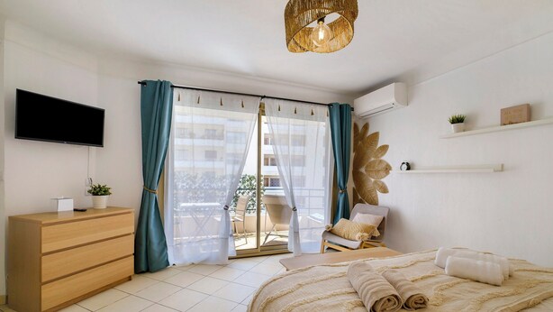 Ein Schlafzimmer mit Bett, Schminktisch, Fernseher, einem Balkon mit Vorhängen und einem an der Wand befestigten Regal mit Pflanze.