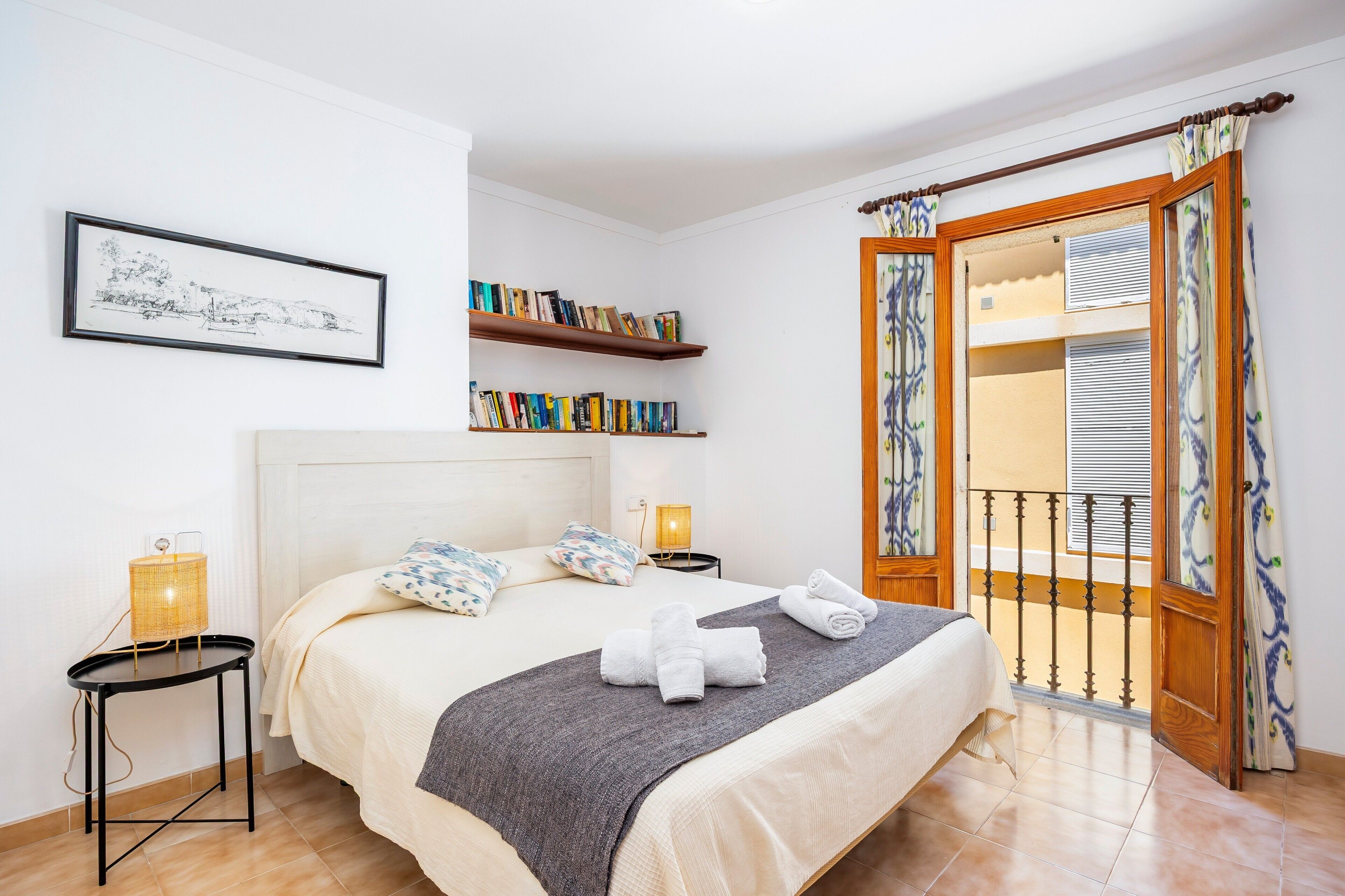 A neatly arranged bedroom with a bed, bedside lamps, a bookshelf, and a balcony with curtains.